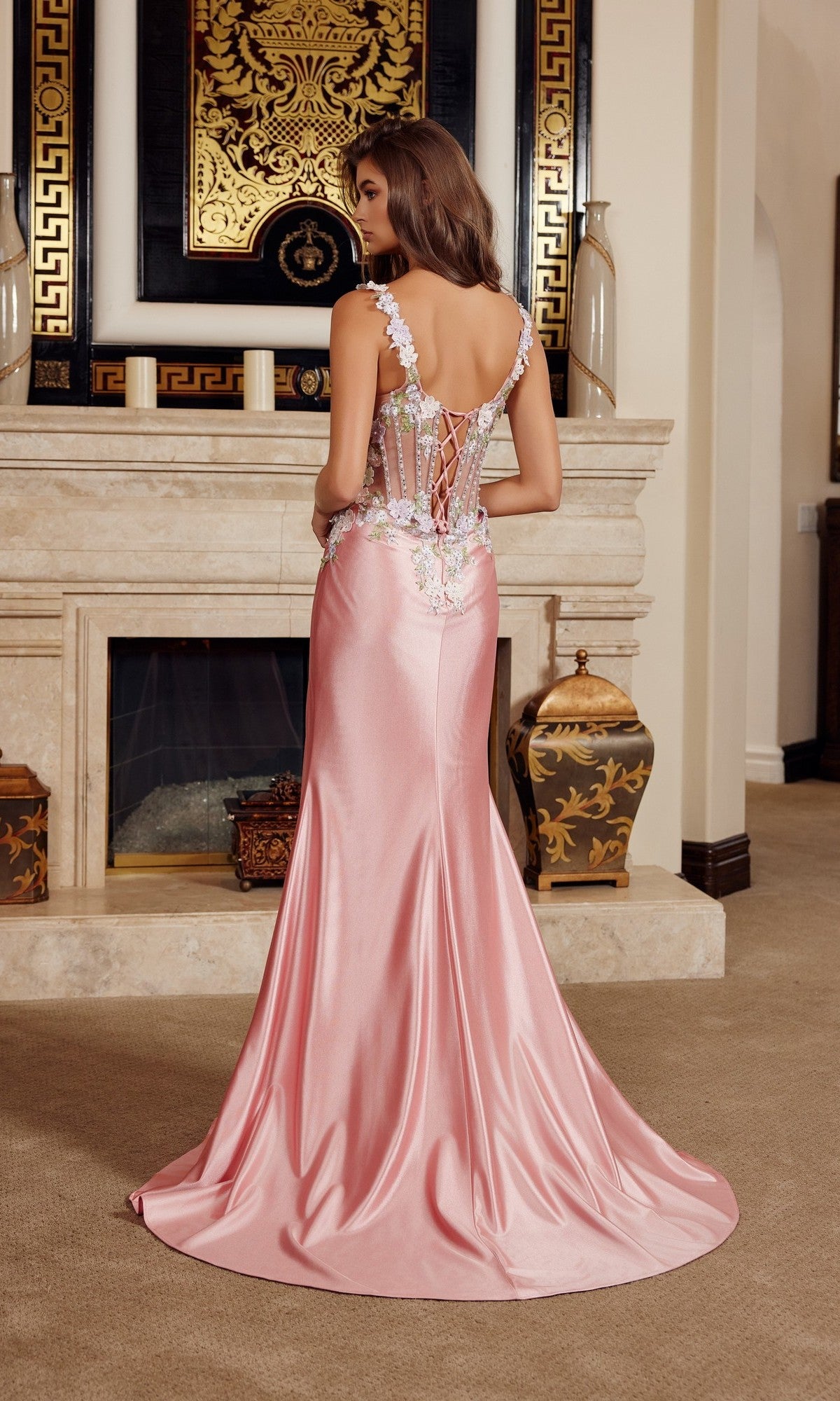 Wearing the Nox Anabel M1755, a woman stands before an ornate fireplace in a sleeveless long prom dress with a floor-length satin skirt and lace-up back, facing away from the camera.
